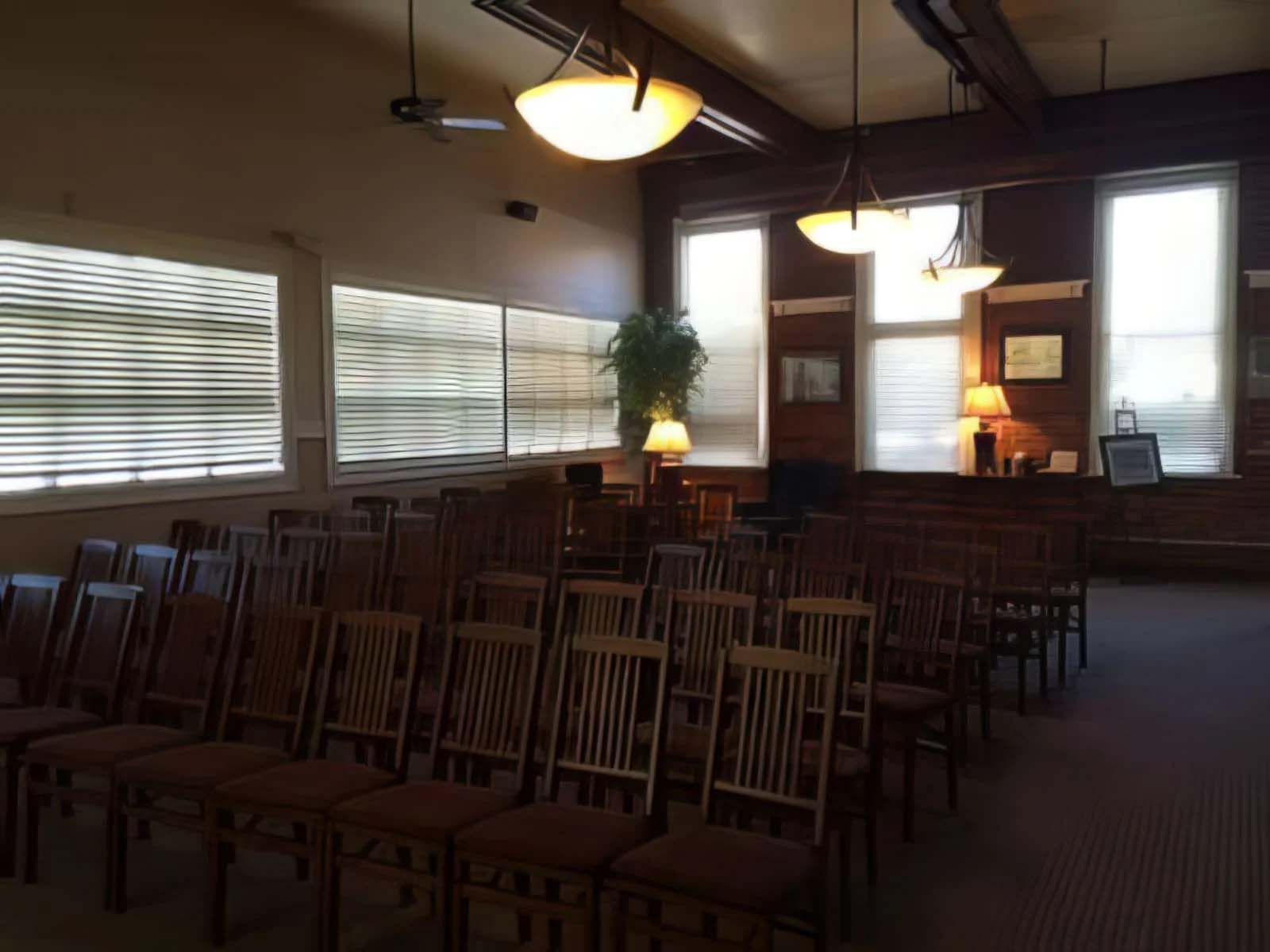 Rows of wooden chairs face a small stage with windows and lamps in a dimly lit room.