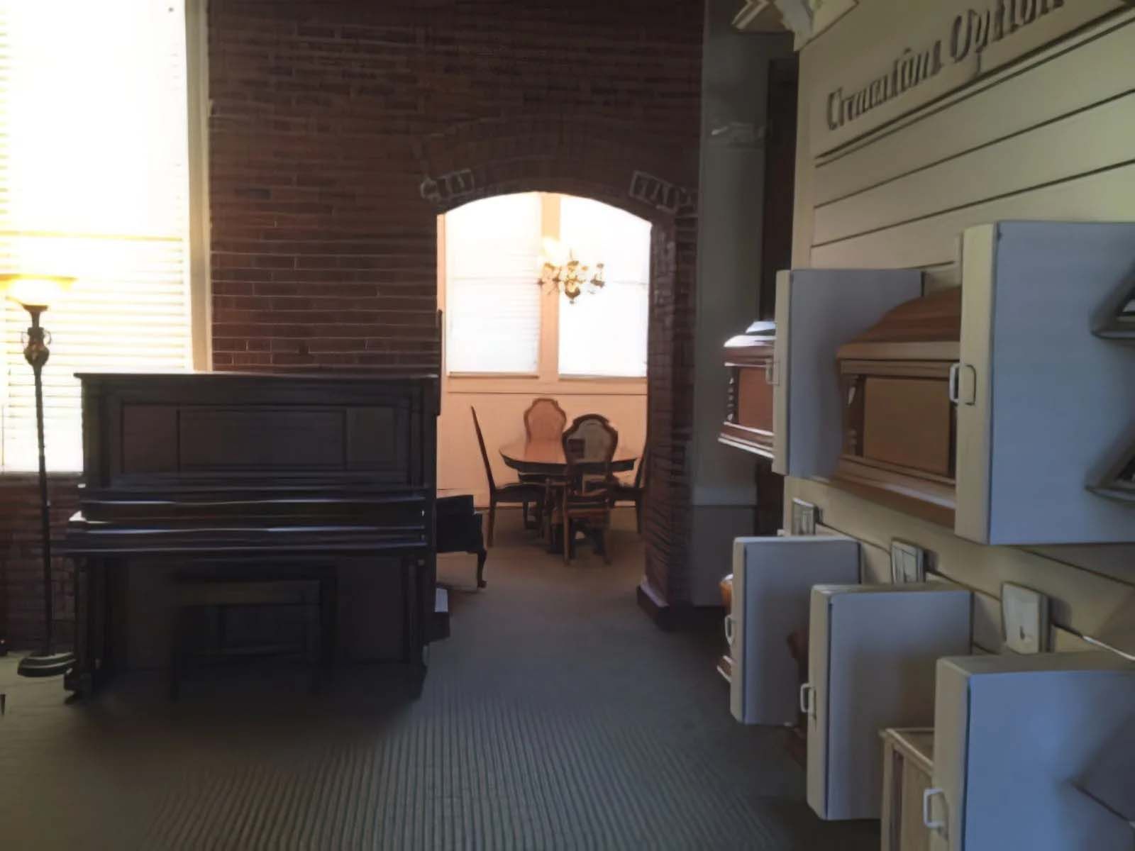 Funeral home interior with a piano, brick wall, archway leading to a table and chairs, and cremation options displayed on the right.