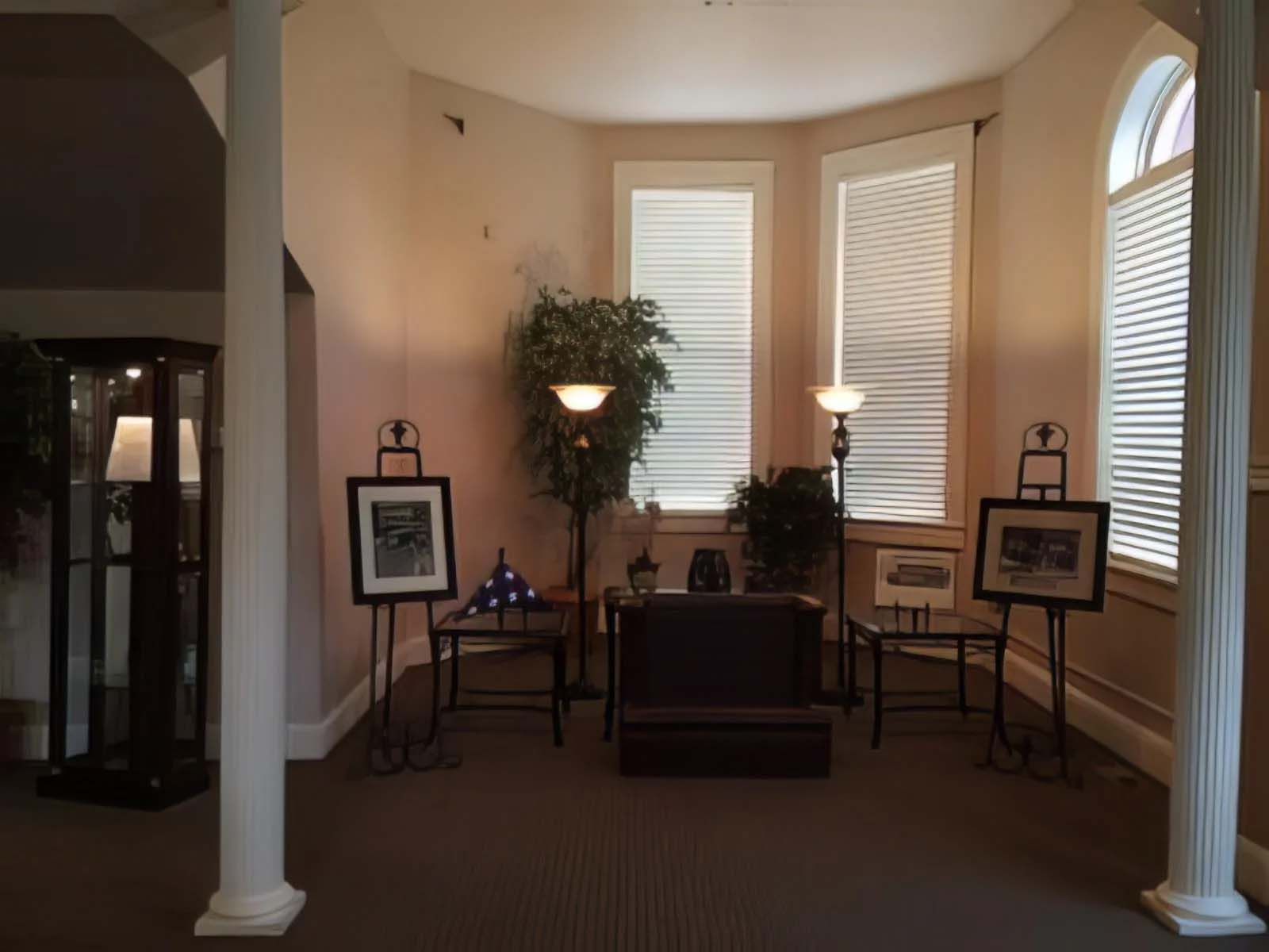 A memorial room with photos on easels, an American flag, and a seating area. The space has large windows, columns, and soft lighting.