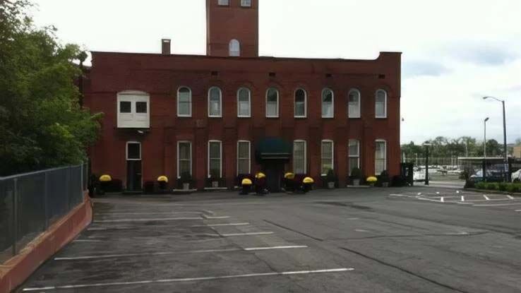 Red brick building with arched windows, a tower, and a small balcony. A paved parking area is in front.