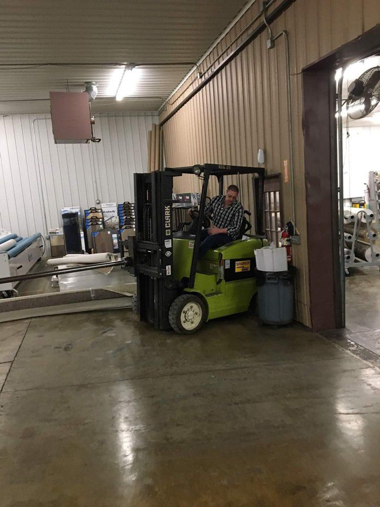 Forklift driver in a warehouse, beside a wall and barrels, with machinery and tables in the background.