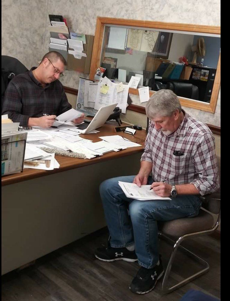 Two people seated at a desk in an office, writing paperwork amid stacks of documents.