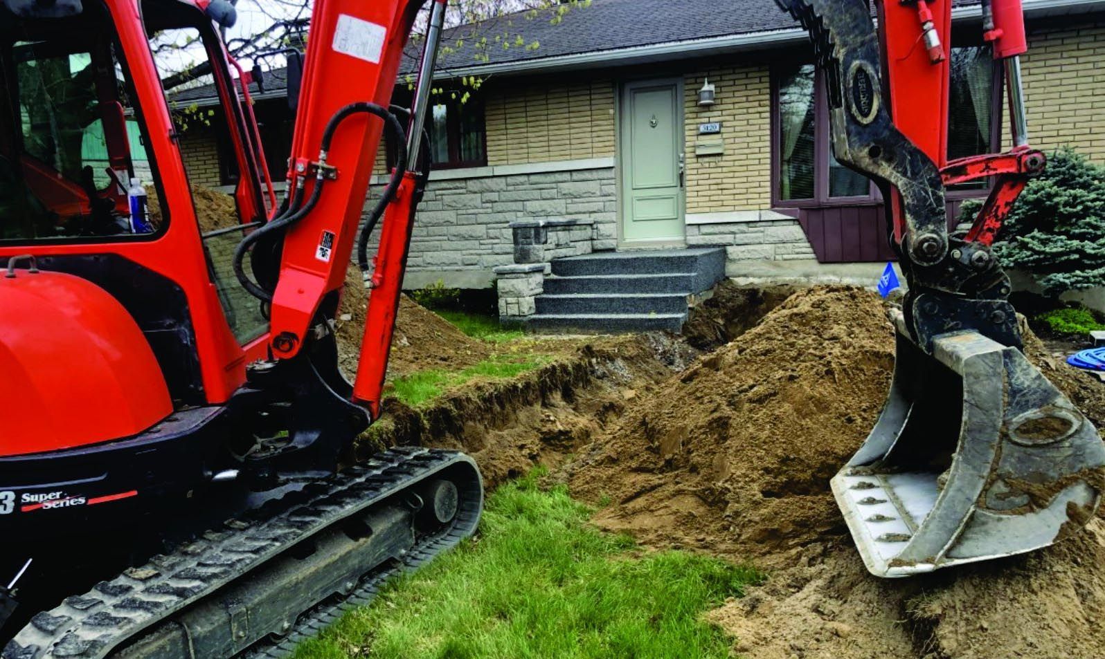 Une excavatrice rouge creuse un trou devant une maison.
