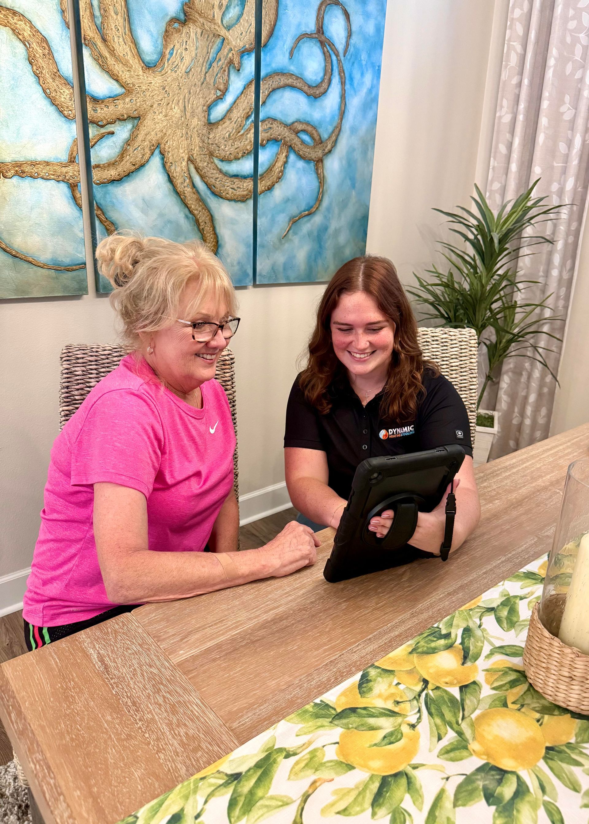 Woman in pink shirt and another person looking at a tablet at a table with an octopus painting in the background.