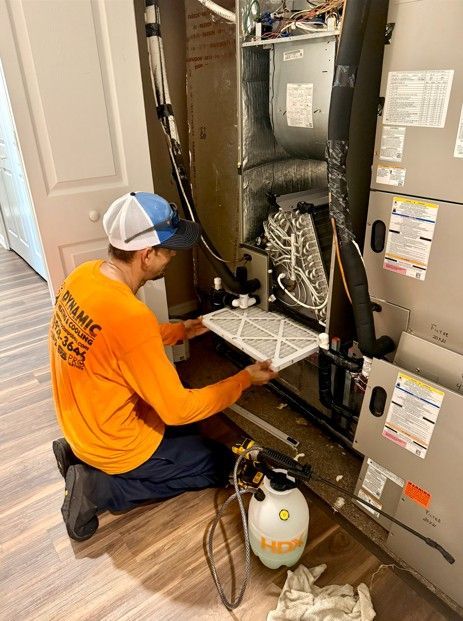 HVAC technician replacing an air filter inside a unit, wearing an orange shirt and cap. A sprayer bottle is on the floor.