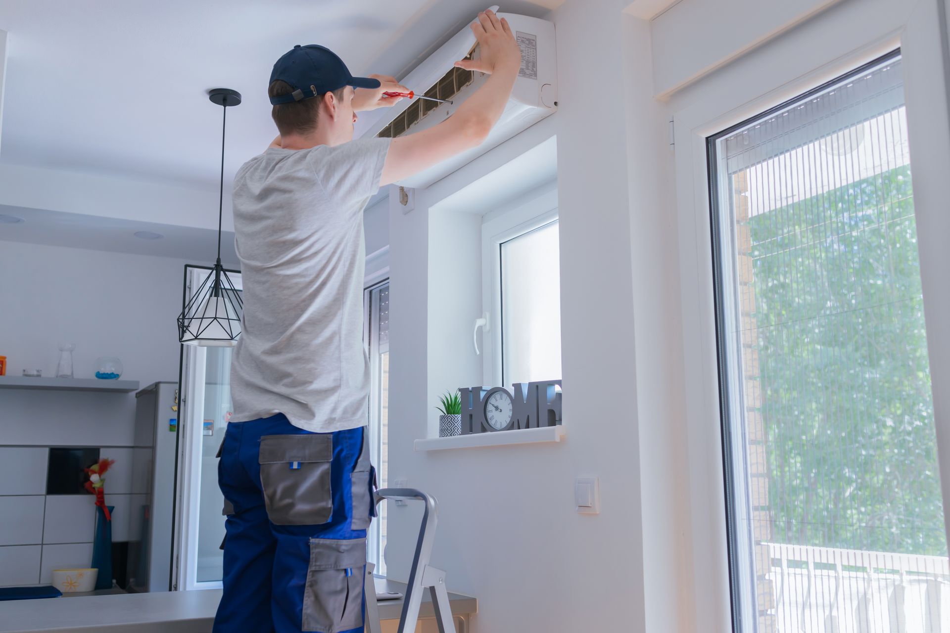 Man on ladder fixing an air conditioner indoors near a window.