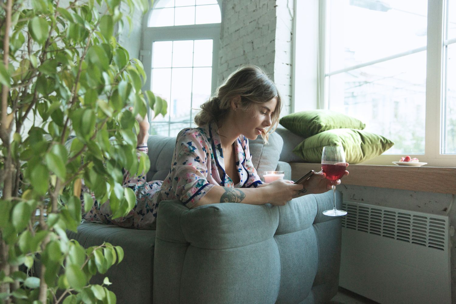 A woman is sitting on a couch holding a glass of wine and looking at her phone.