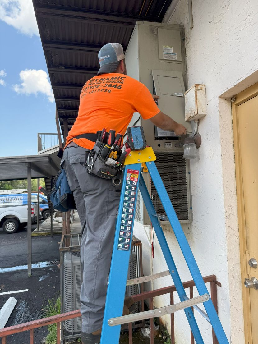 An electrician in an orange shirt works on an air conditioning unit outside a building while standing on a blue ladder.