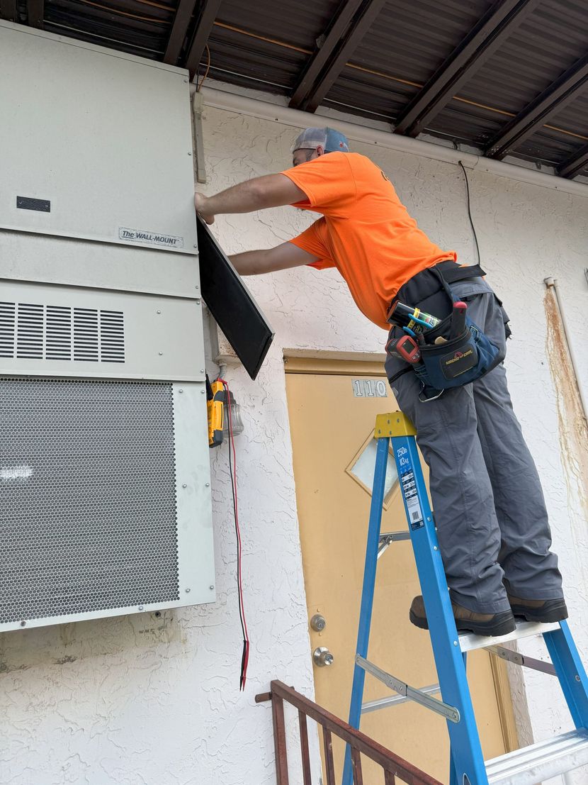 A person in an orange shirt and gray pants installs a dark panel on an air conditioning unit, using a ladder outside a building.