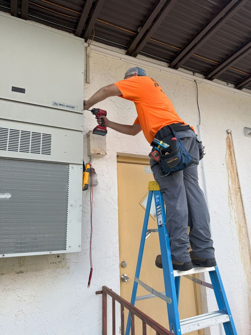 Man in orange shirt uses a drill while standing on a blue ladder, working on an electrical box attached to a building.