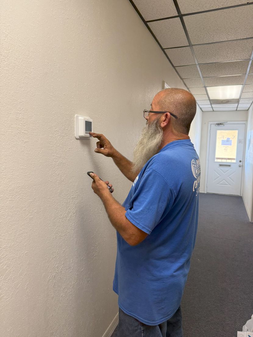 Man with a long beard adjusts a thermostat on a textured wall in an office hallway. He wears a blue shirt and glasses.