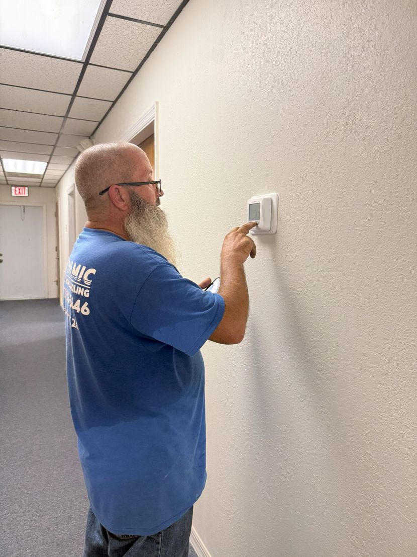 Man adjusting thermostat on a white wall in a hallway. He wears a blue shirt and glasses.