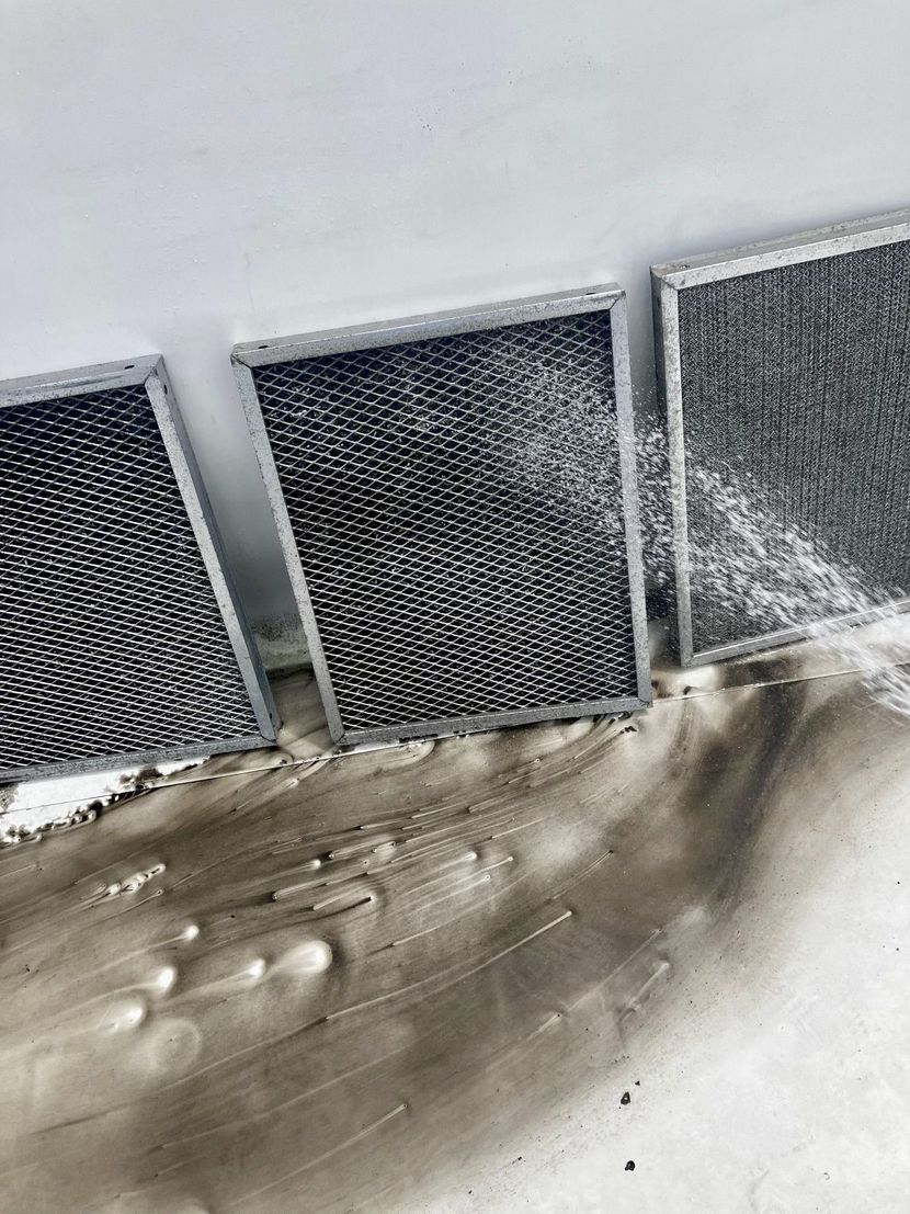 Three dirty metallic mesh air filters resting on a floor covered in black grime, against a white wall.