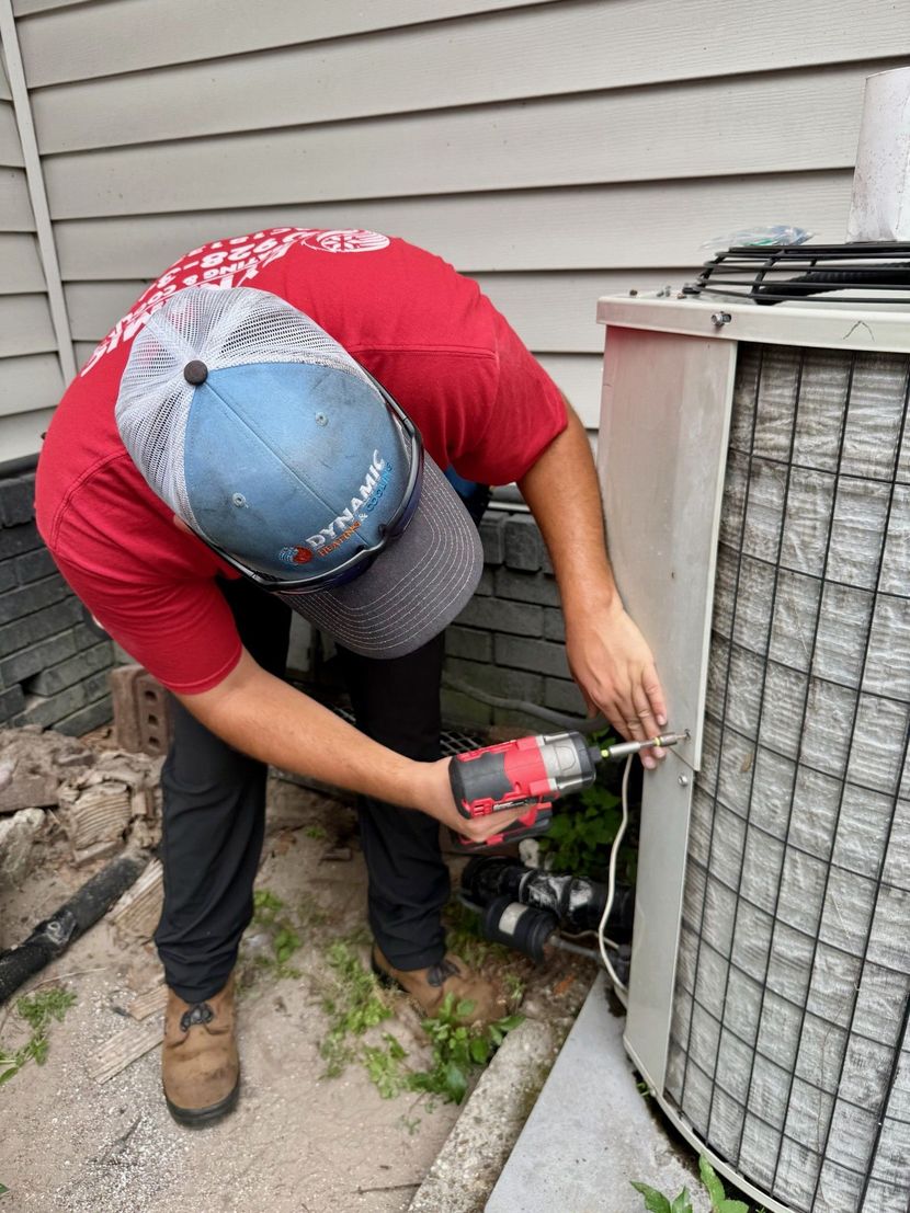 A person wearing a red shirt and hat uses a drill to work on an air conditioning unit outside a building.