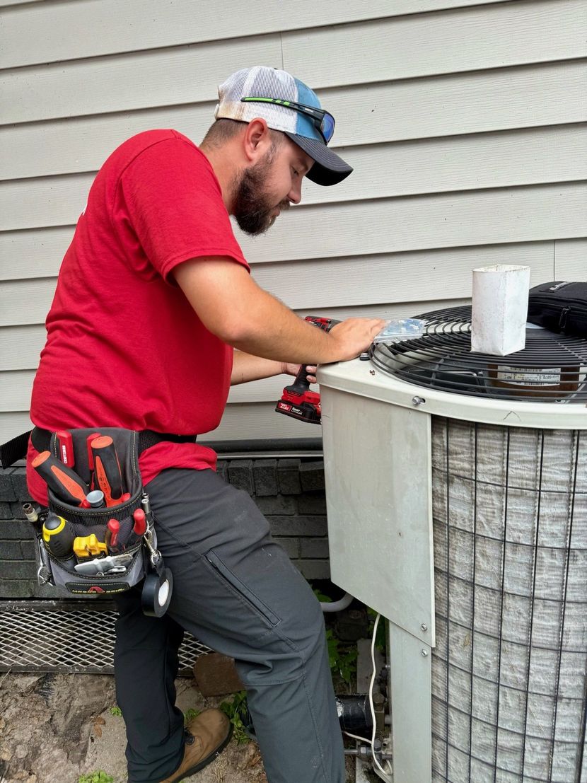 HVAC technician in a red shirt repairs an air conditioning unit outside a house. He's using a drill and wearing a tool belt.