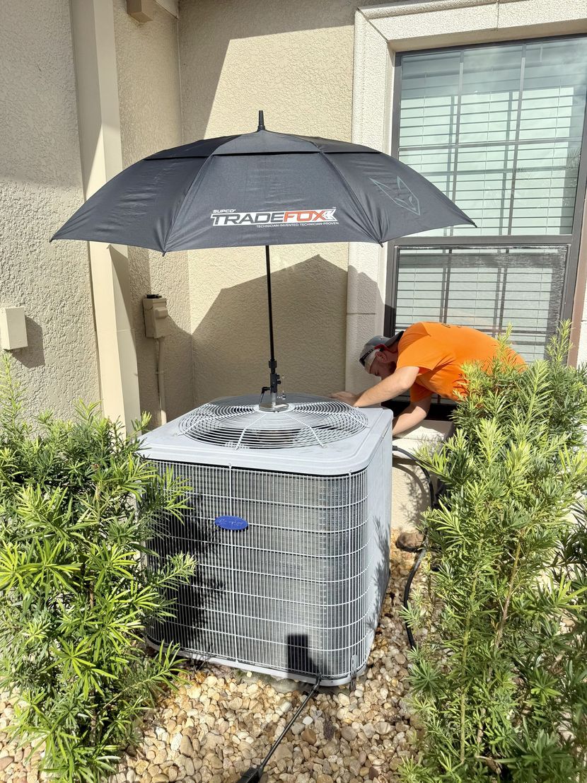 A person in an orange shirt working on an air conditioner unit, shaded by an umbrella. The unit is outside near a window and bushes.