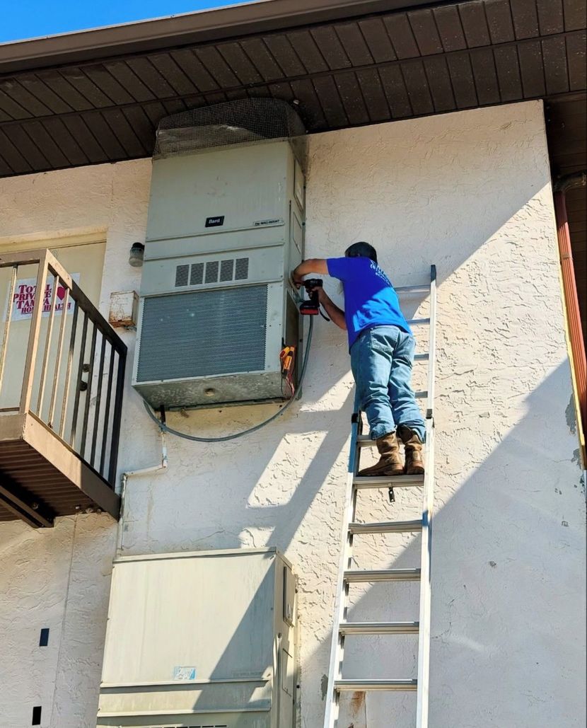 A person in blue shirt and jeans on a ladder working on an air conditioning unit attached to a building's wall.
