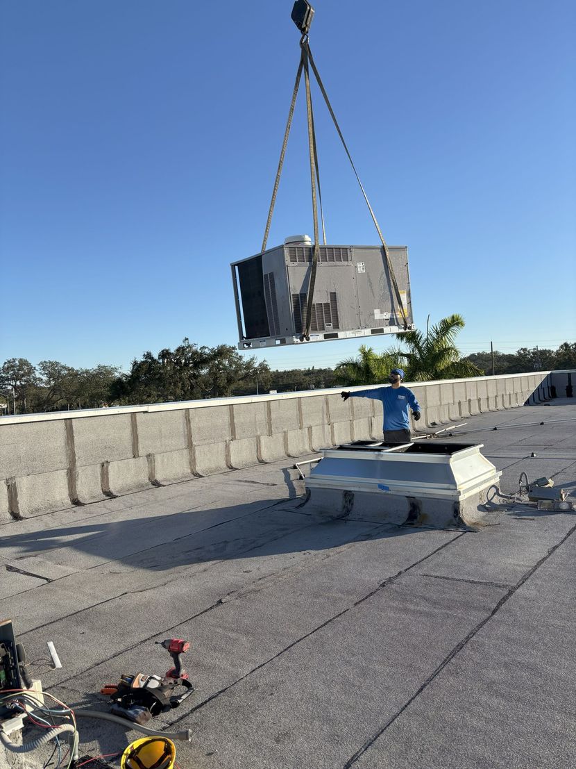 A crane lifts a large HVAC unit over a flat rooftop. Blue sky overhead.