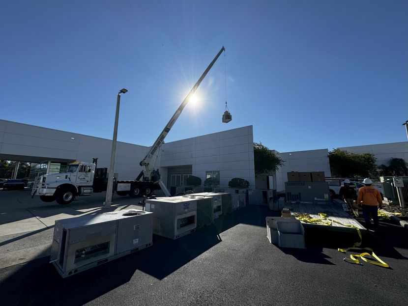 A crane lifting equipment near a white building under a bright sun. Construction workers are visible on the ground.