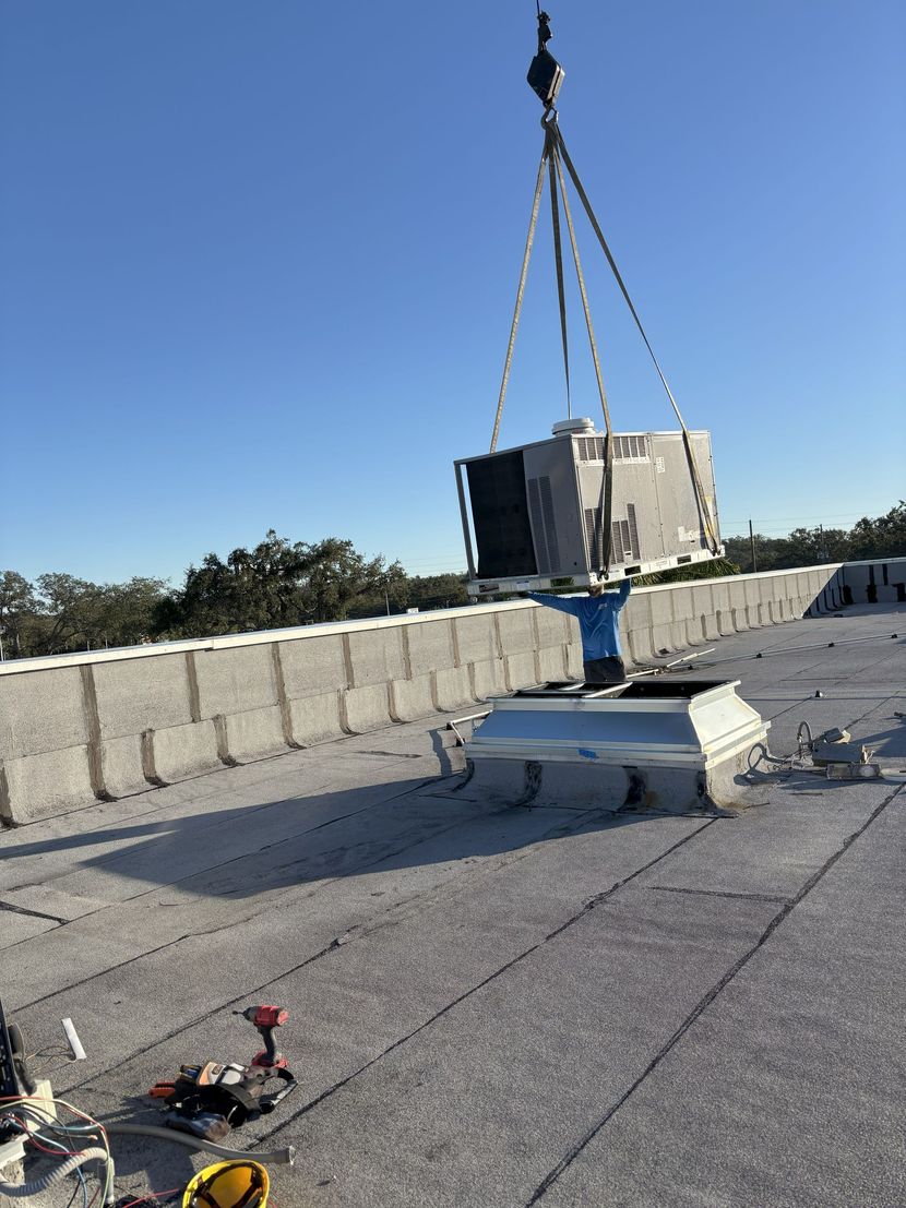 A crane lifts an HVAC unit onto a rooftop, with the new unit above a vent opening. Bright sky, sunny day.
