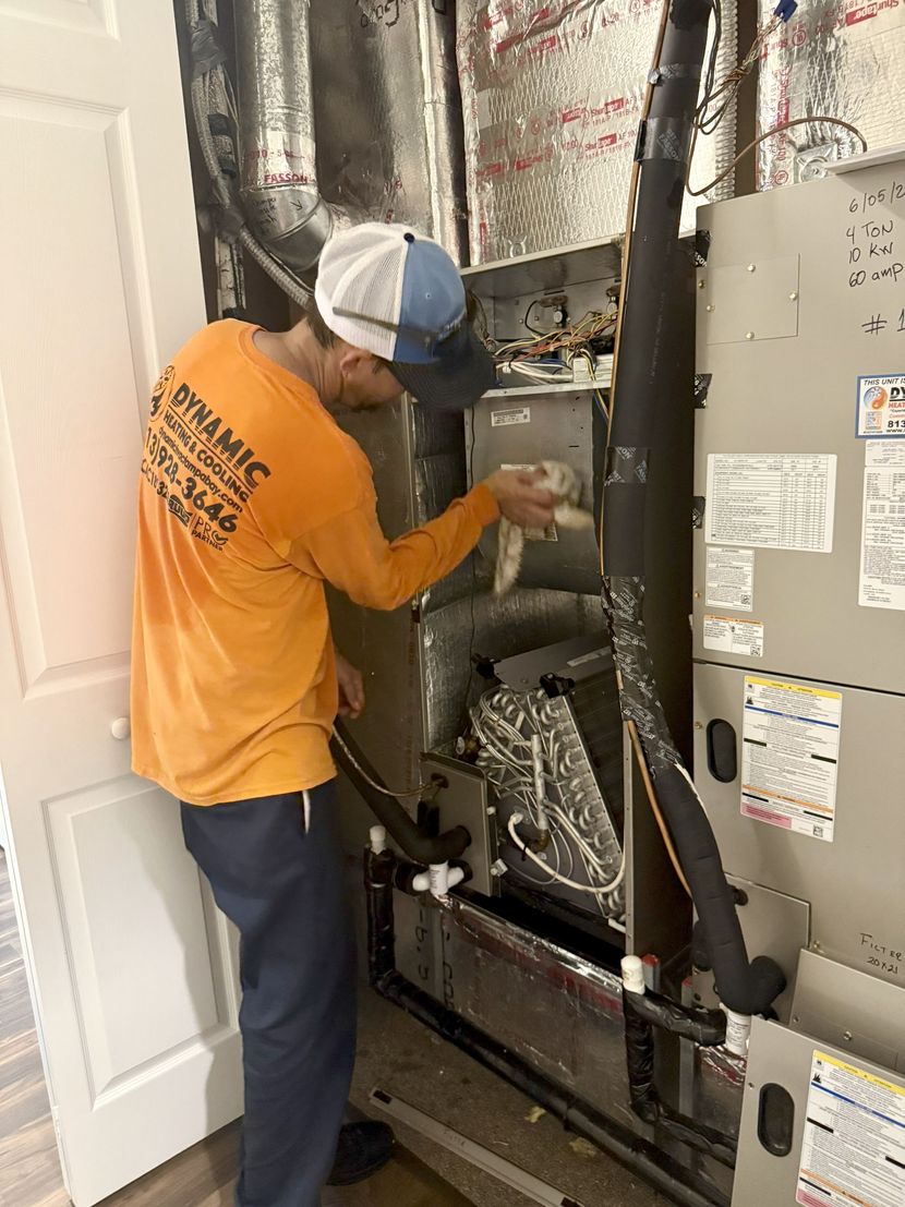 A person in an orange shirt and blue hat works on an HVAC unit. Indoors, the unit is open.