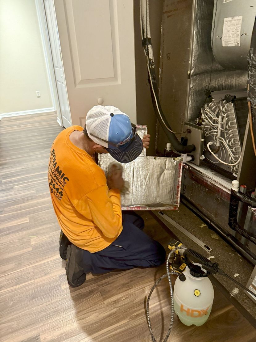 HVAC technician, wearing orange shirt and blue hat, working on an air conditioning unit. Kneeling on a hardwood floor.