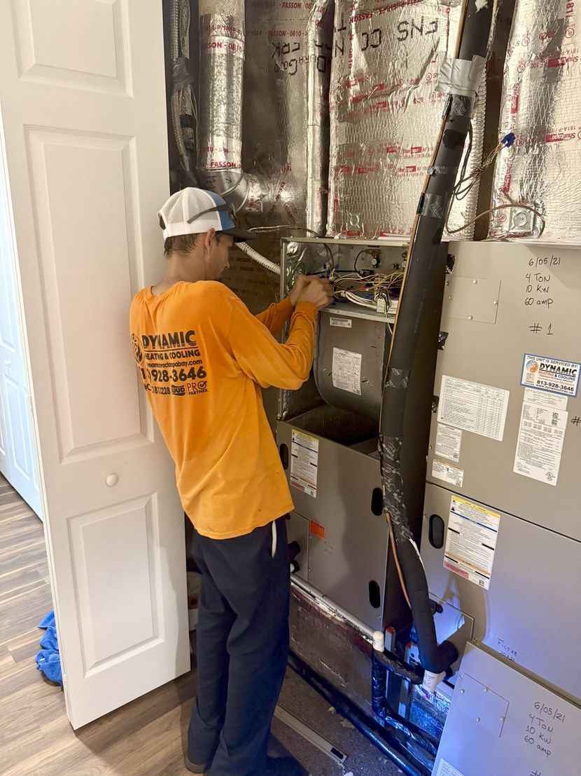 HVAC technician in an orange shirt is working on an air conditioning unit in a wall. The man is inside a utility closet.