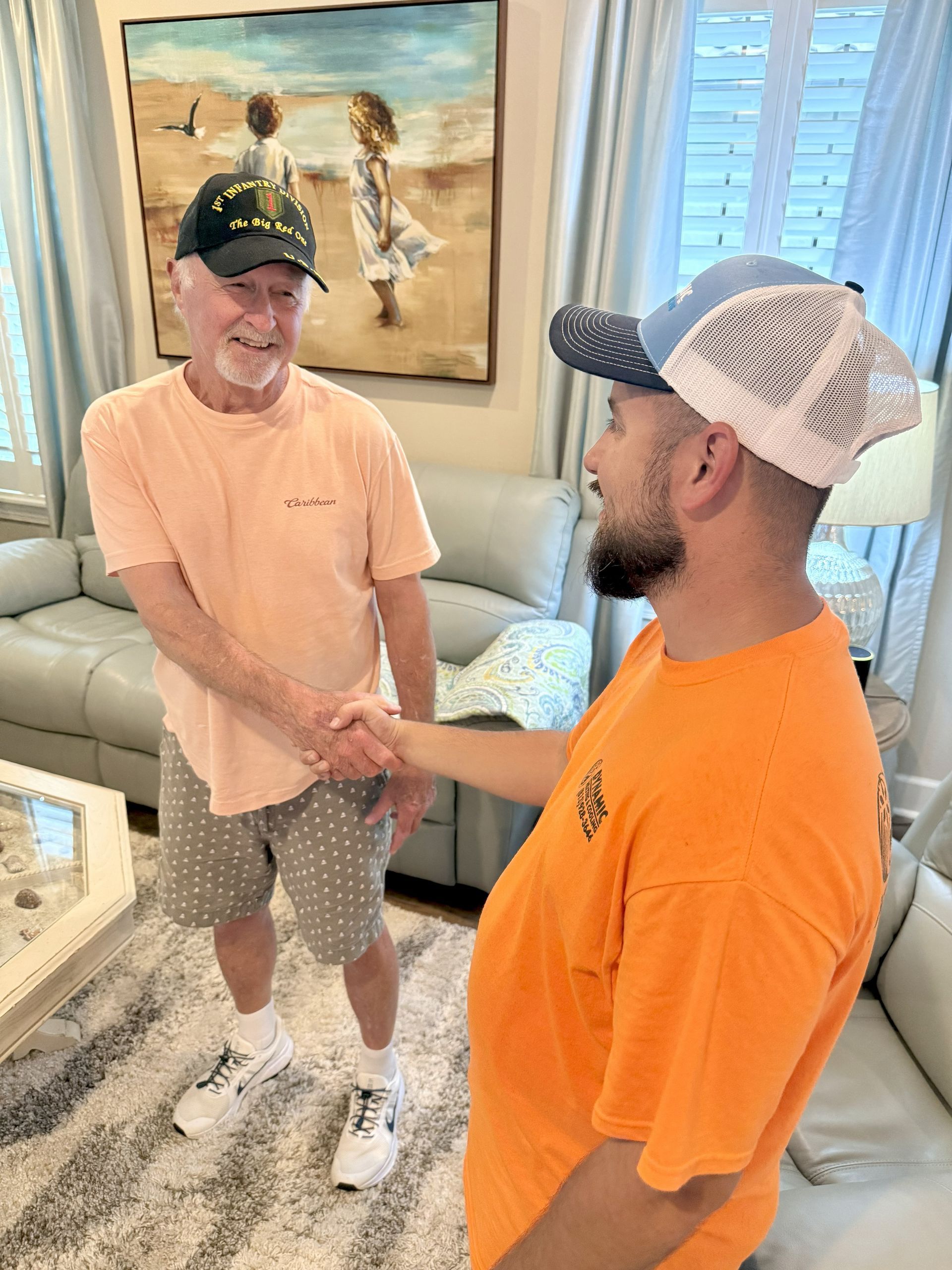 Two men shaking hands indoors. One wears a peach shirt, hat, and shorts. The other man wears an orange shirt and a blue hat.