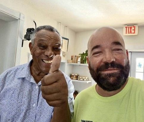 Two men smiling, one giving a thumbs up. One man is Black, in a blue shirt; the other is white with a beard, in a yellow shirt. Inside a store.