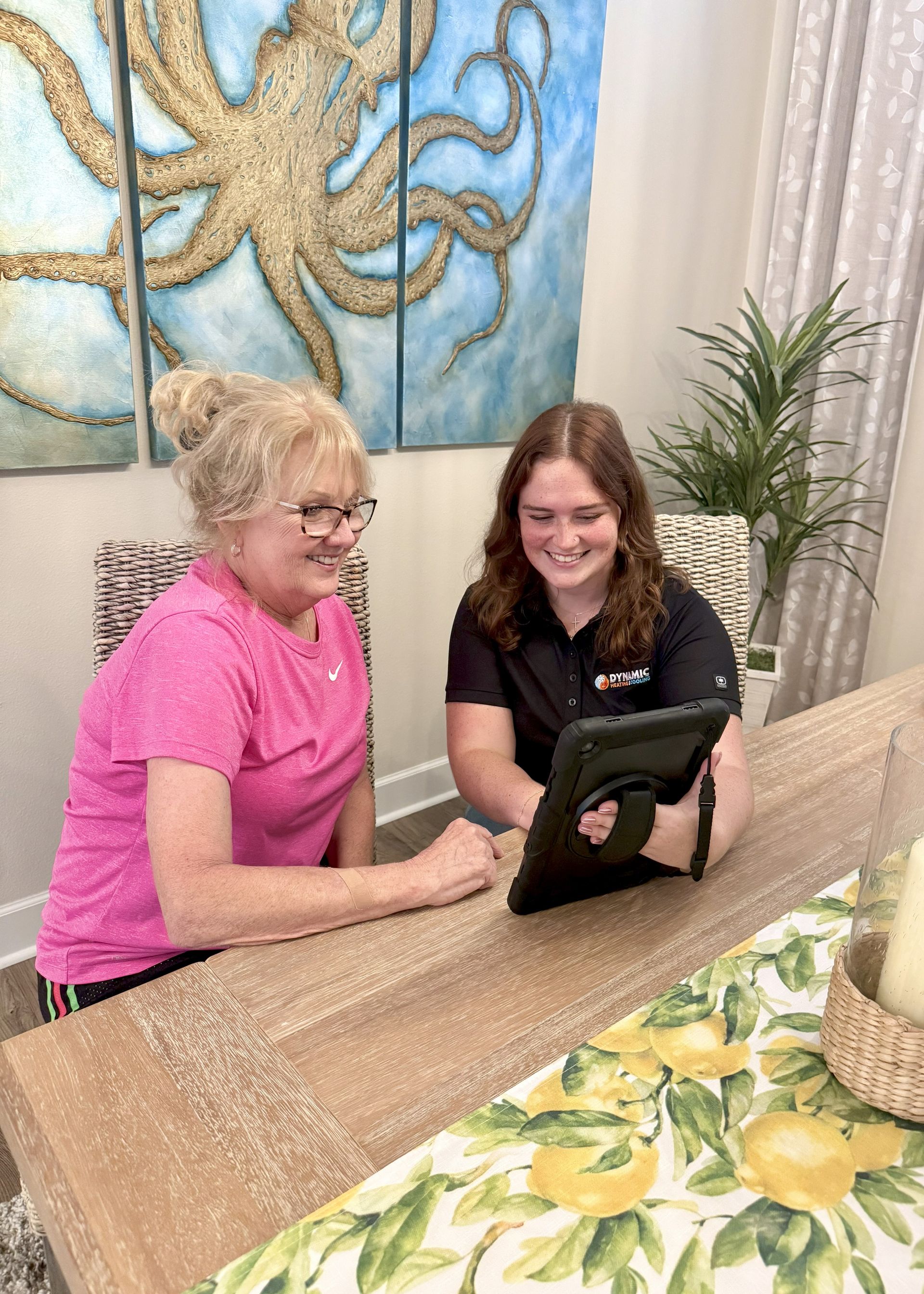 Two women looking at a tablet together at a dining table. One older woman wears a pink shirt and glasses; the other wears a black polo shirt.