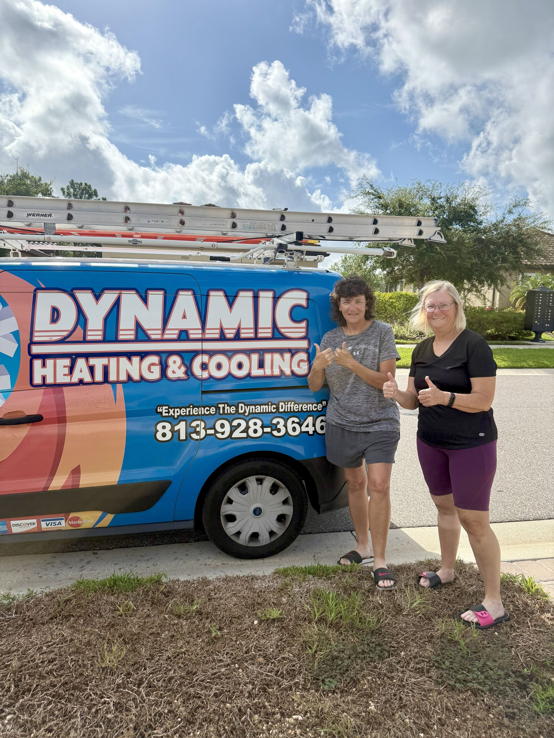 Two women give thumbs up next to a Dynamic Heating & Cooling van under a sunny sky.