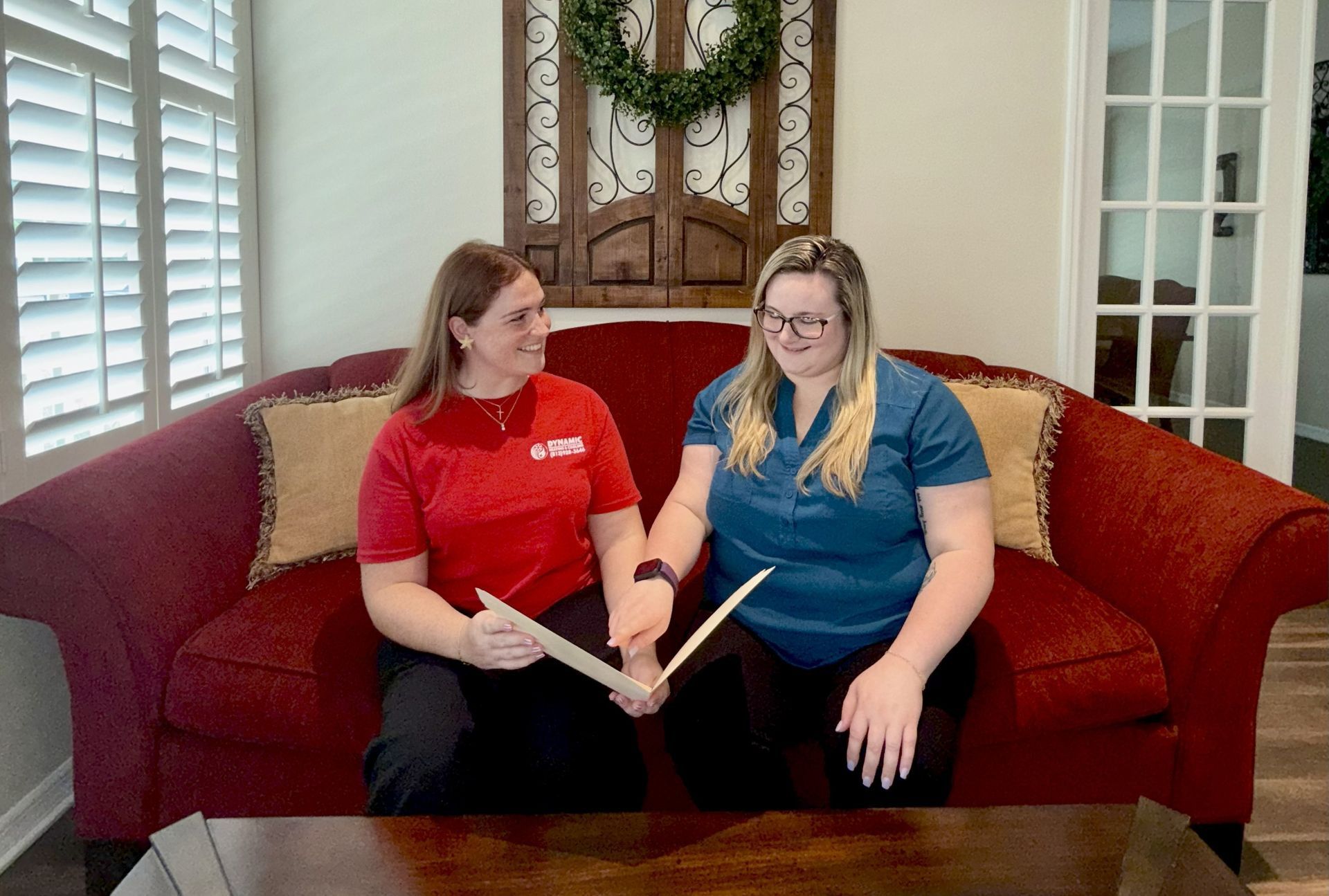 Two women, one in red and one in blue, seated on a red couch, holding and examining bamboo sticks. They are smiling indoors with a wreath in the background.