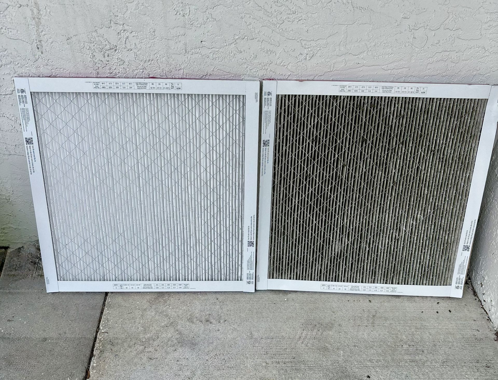 Two air filters, one clean, one dirty, resting on a concrete surface in front of a white wall.