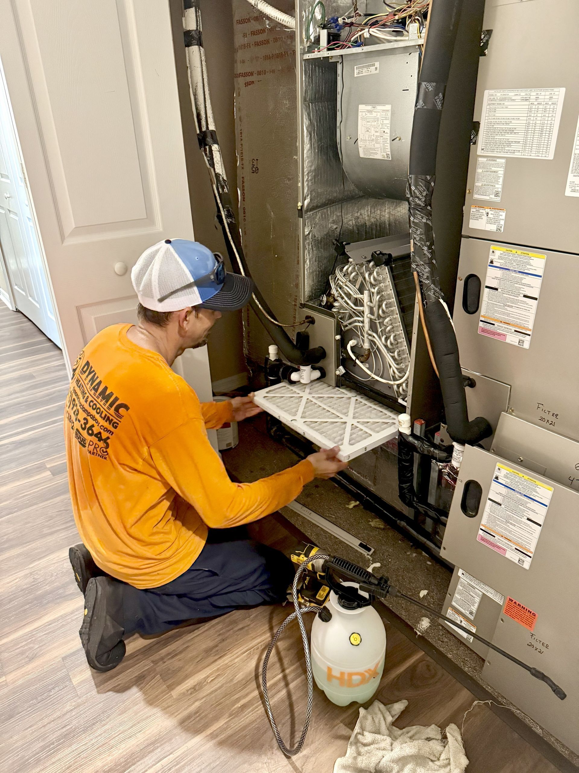 HVAC technician replacing an air filter in a home's air handler unit. He is wearing a hat and orange shirt, kneeling.