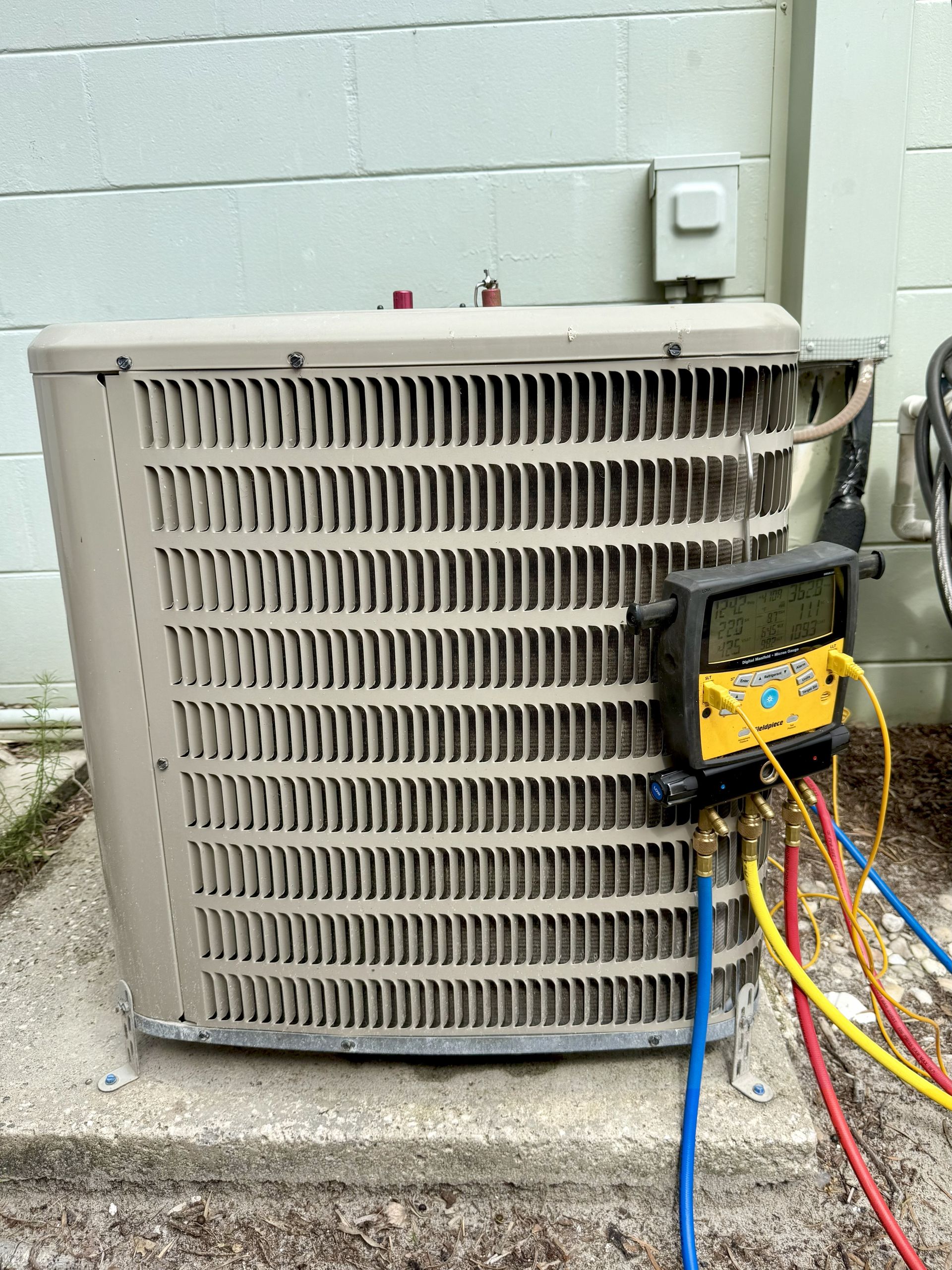 An air conditioning unit with a yellow gauge attached, and colored hoses extending from it. The unit sits on a concrete pad near a light colored building.