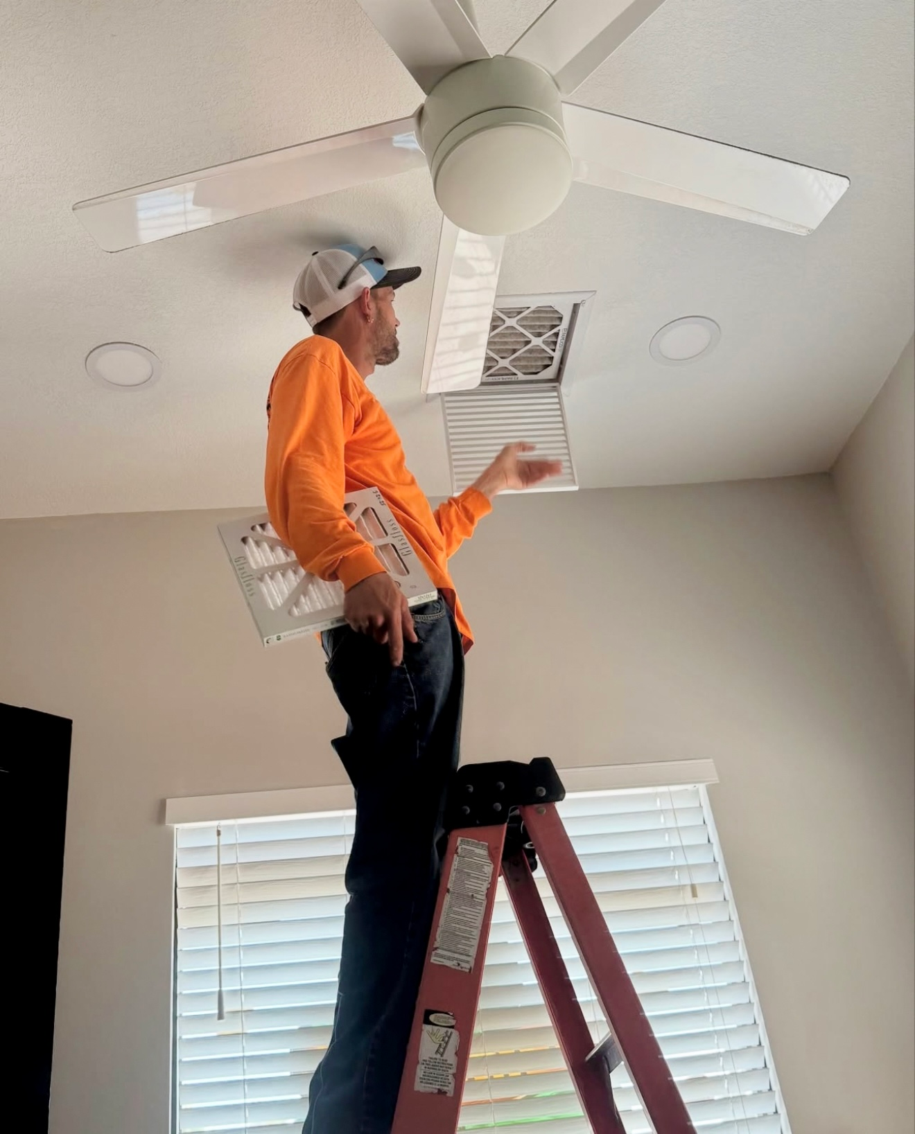 Man on a ladder replacing an air vent filter near a ceiling fan. He wears an orange shirt and cap in a room with a window.