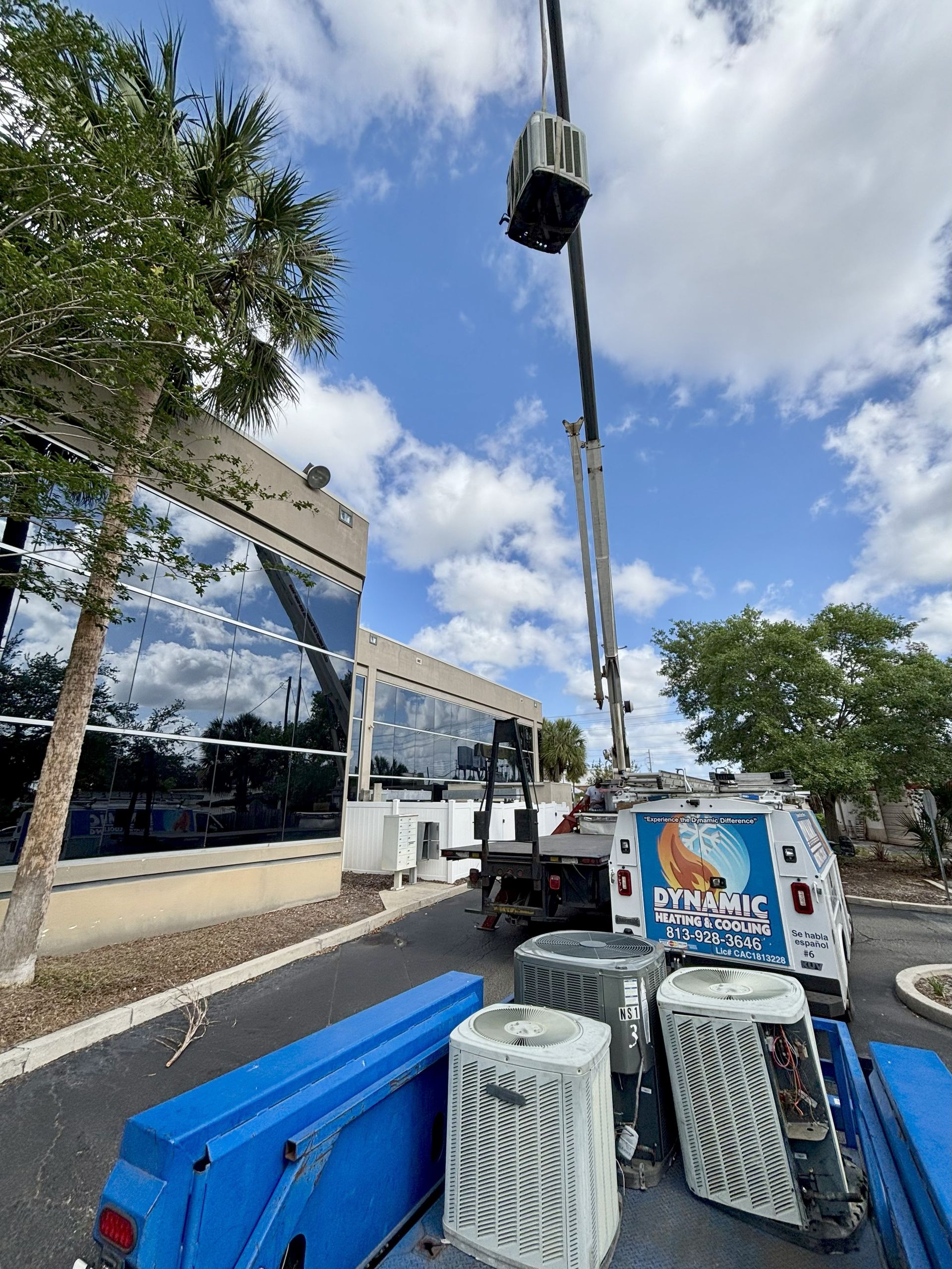 A crane lifts an AC unit towards a building. Several other AC units and blue barriers are on the ground near a service truck on a sunny day.