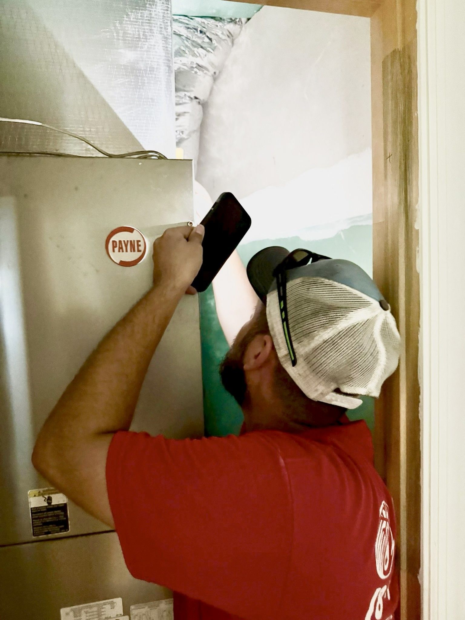 A man in a red shirt examines an HVAC unit, shining a light. He wears a white cap and is in a room with exposed insulation.