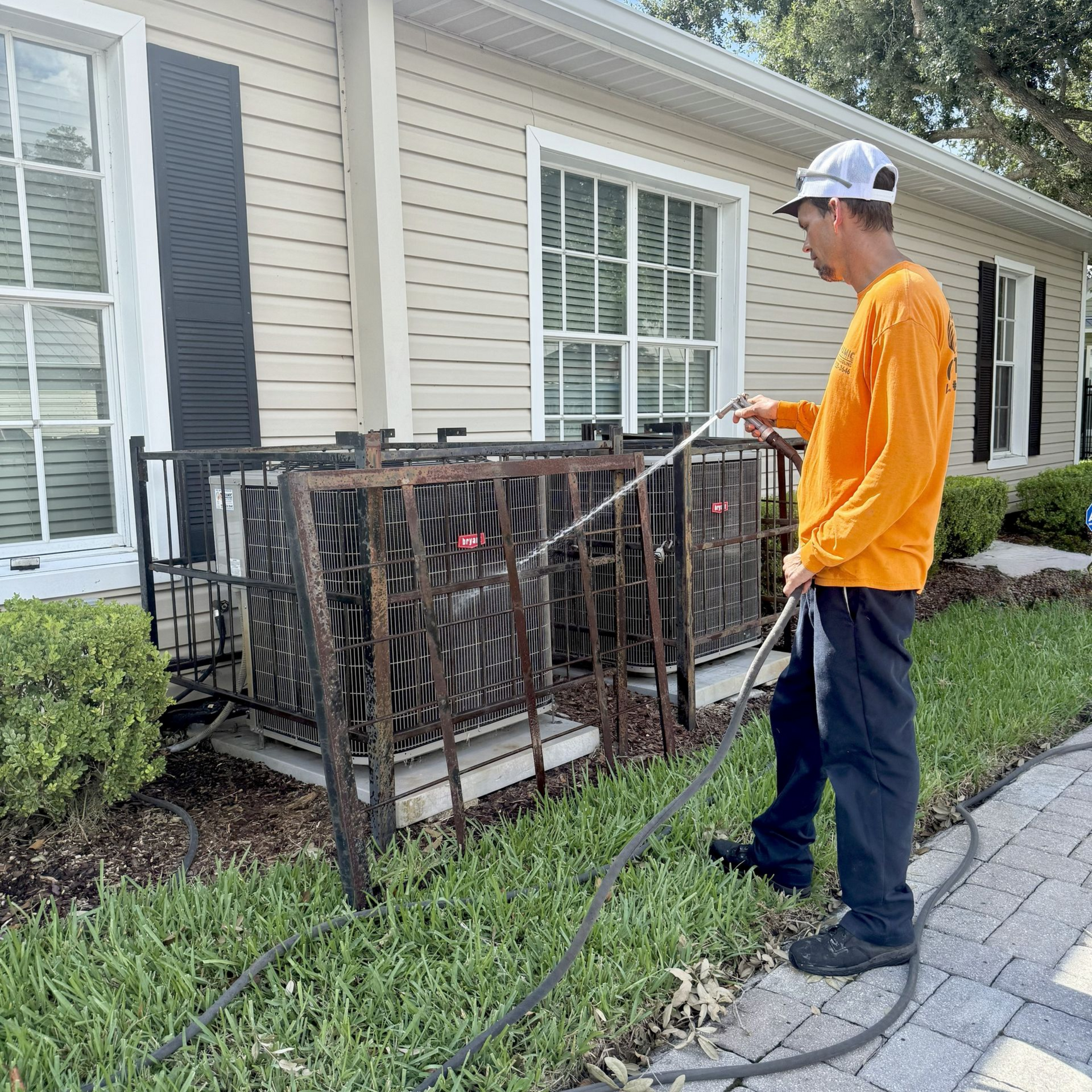 Man in orange shirt sprays an AC unit with a hose, standing near a beige building, bushes, and paved walkway.
