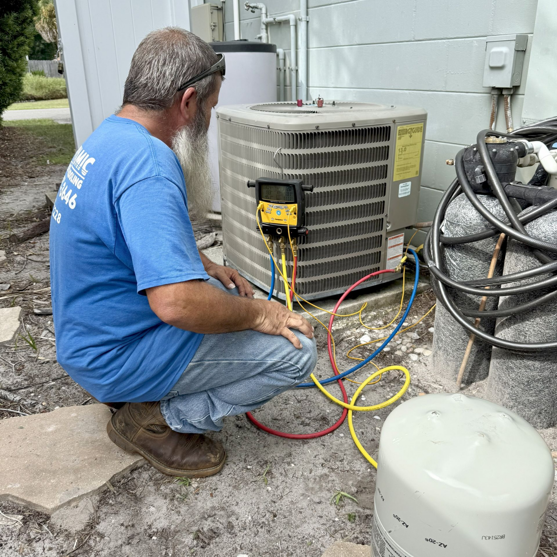 HVAC technician crouches, connecting gauges to an air conditioning unit outside a light-colored building.