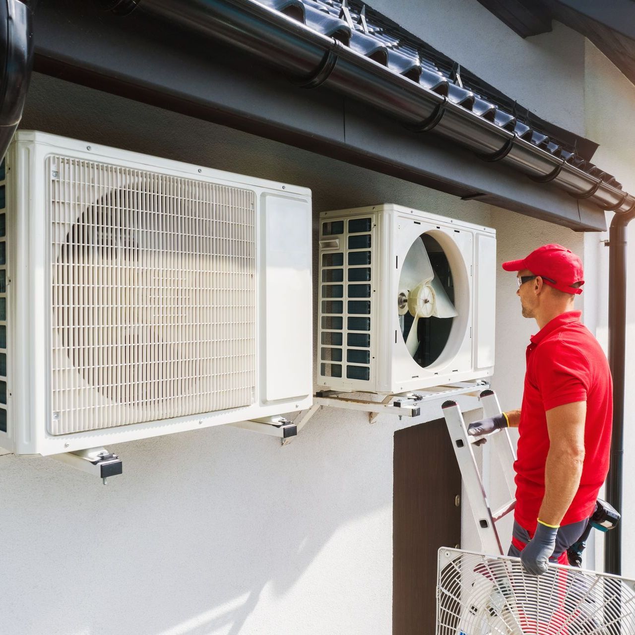 A man is standing on a ladder next to two air conditioners on the side of a building.