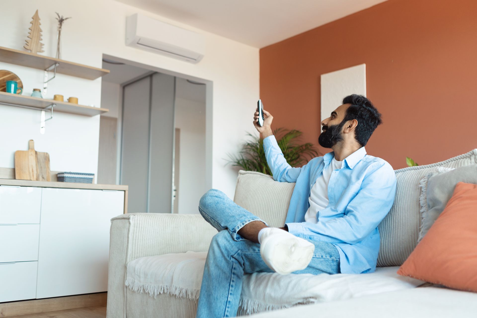 Man in light blue shirt and jeans sits on a sofa, looking at his phone in a living room.