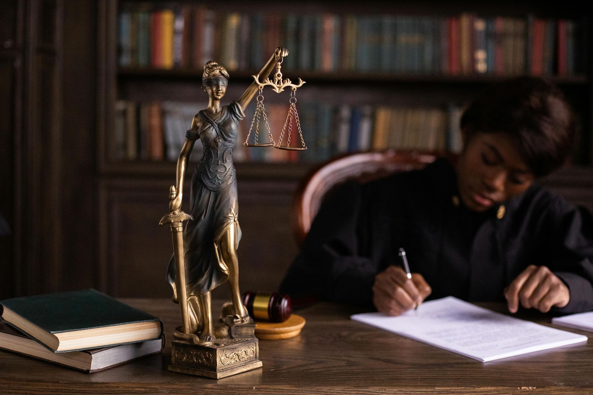 Lady Justice statue beside a person writing at a desk in a library-like room