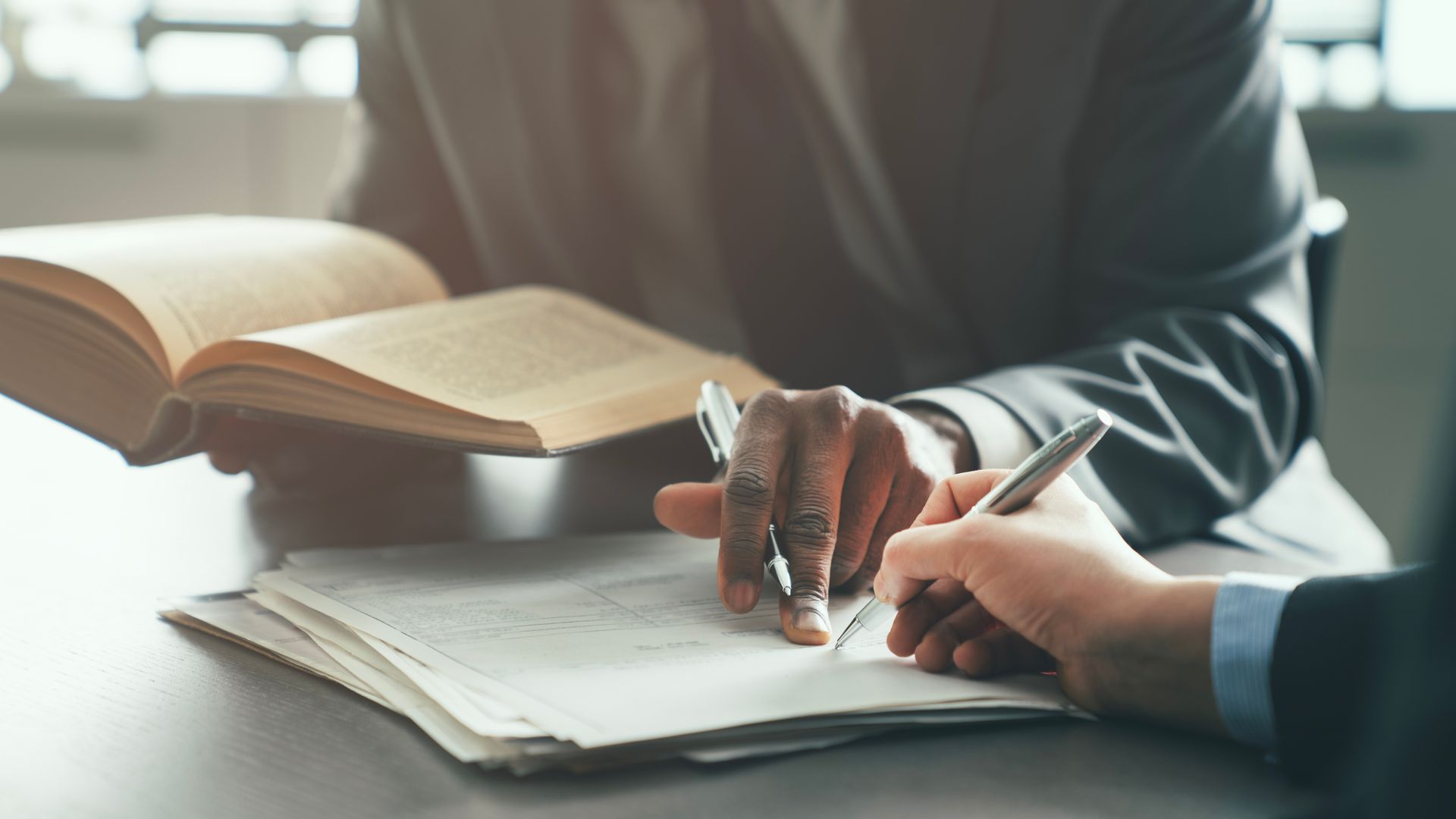 Two people reviewing paperwork at a desk, one holding an open book and the other writing on a form.