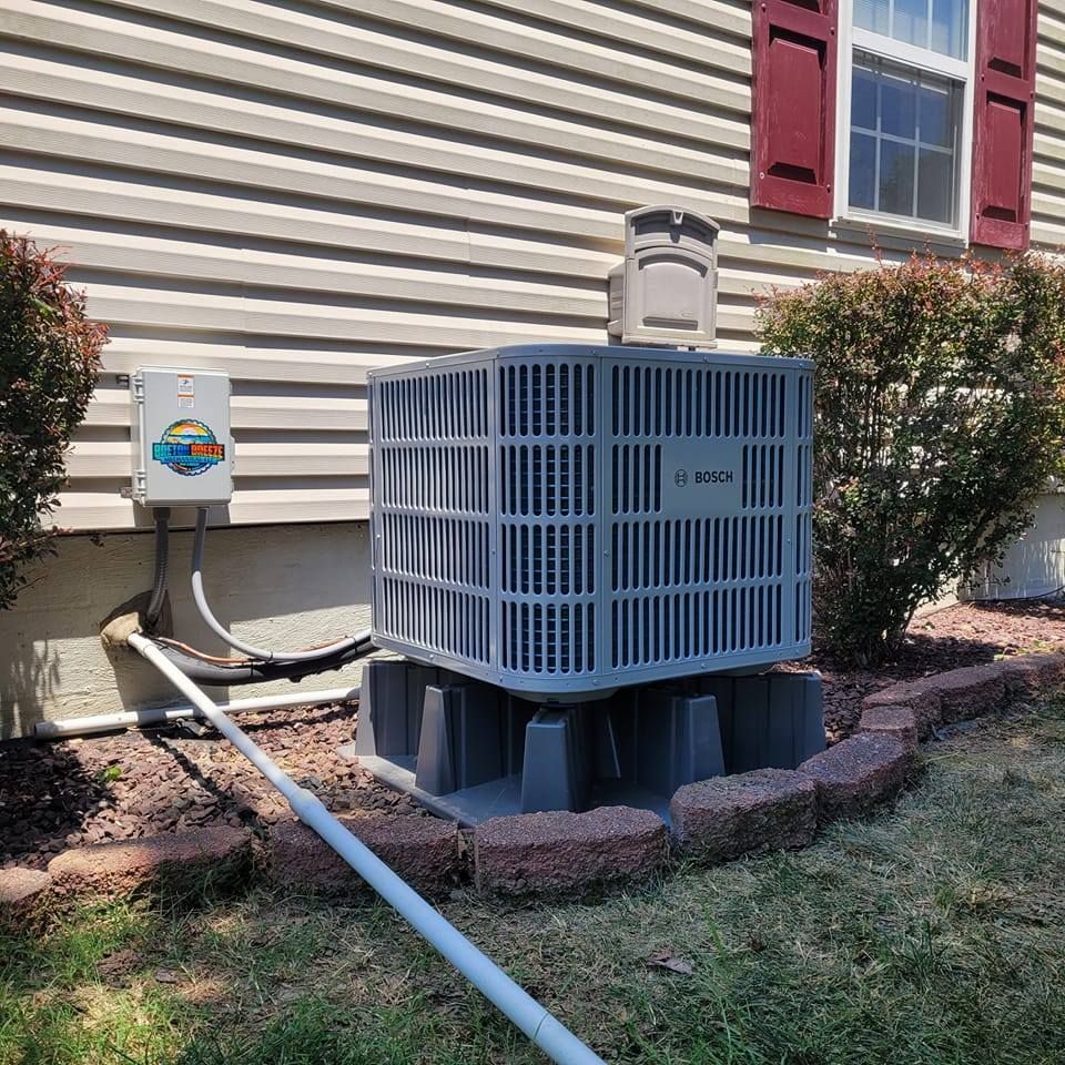 Gray air conditioning unit on a concrete base, next to a home with brown siding and a window.