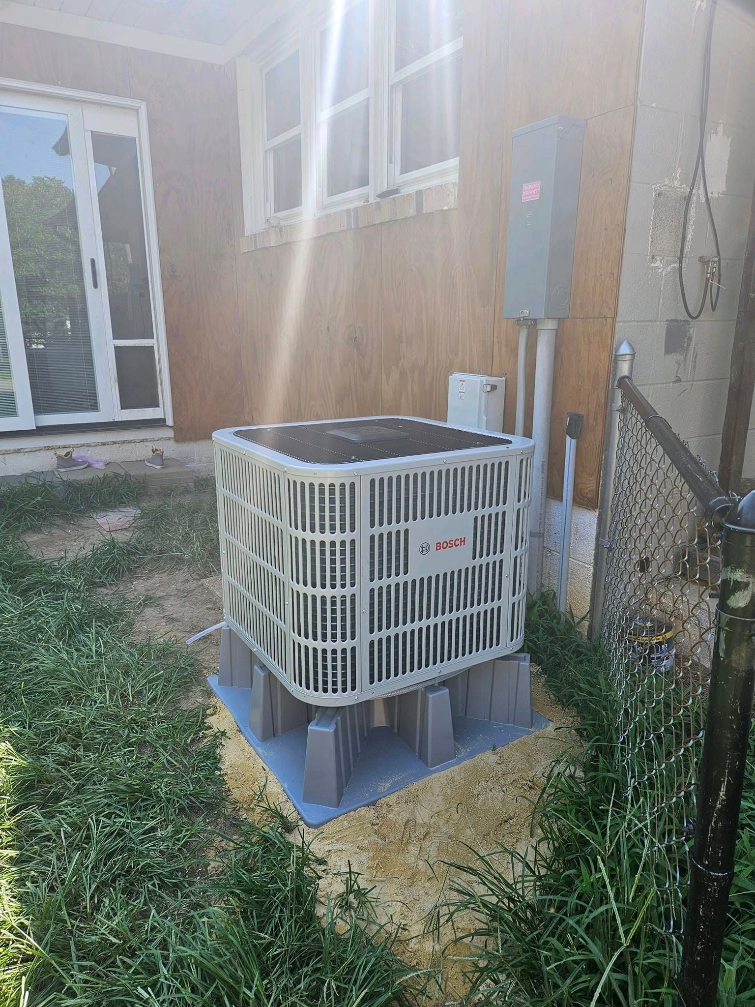 Air conditioner unit outside a building, on a raised platform, with electrical box and overgrown grass.