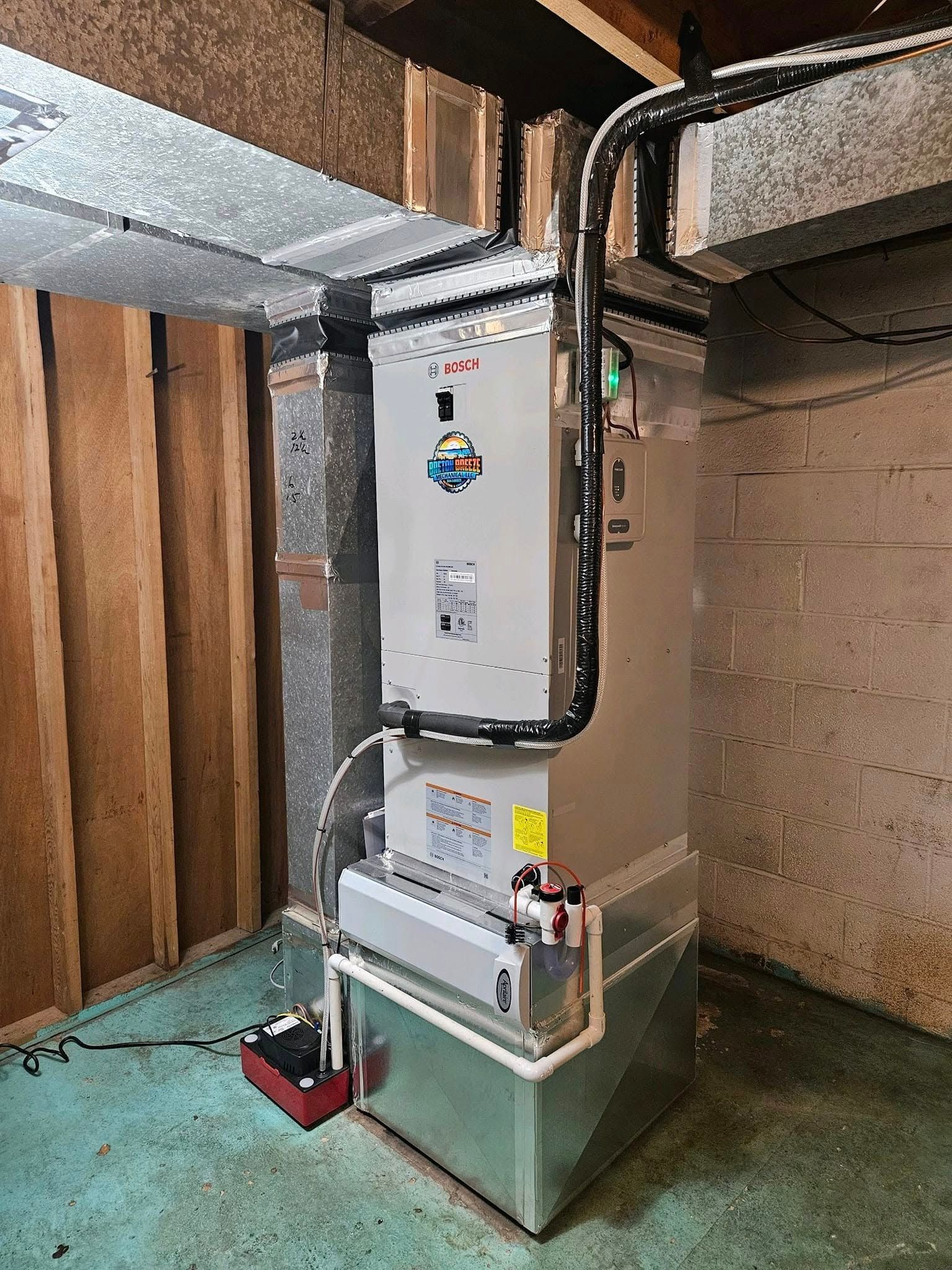 Furnace in a basement, silver and white with black pipe, next to a wooden wall and concrete floor.