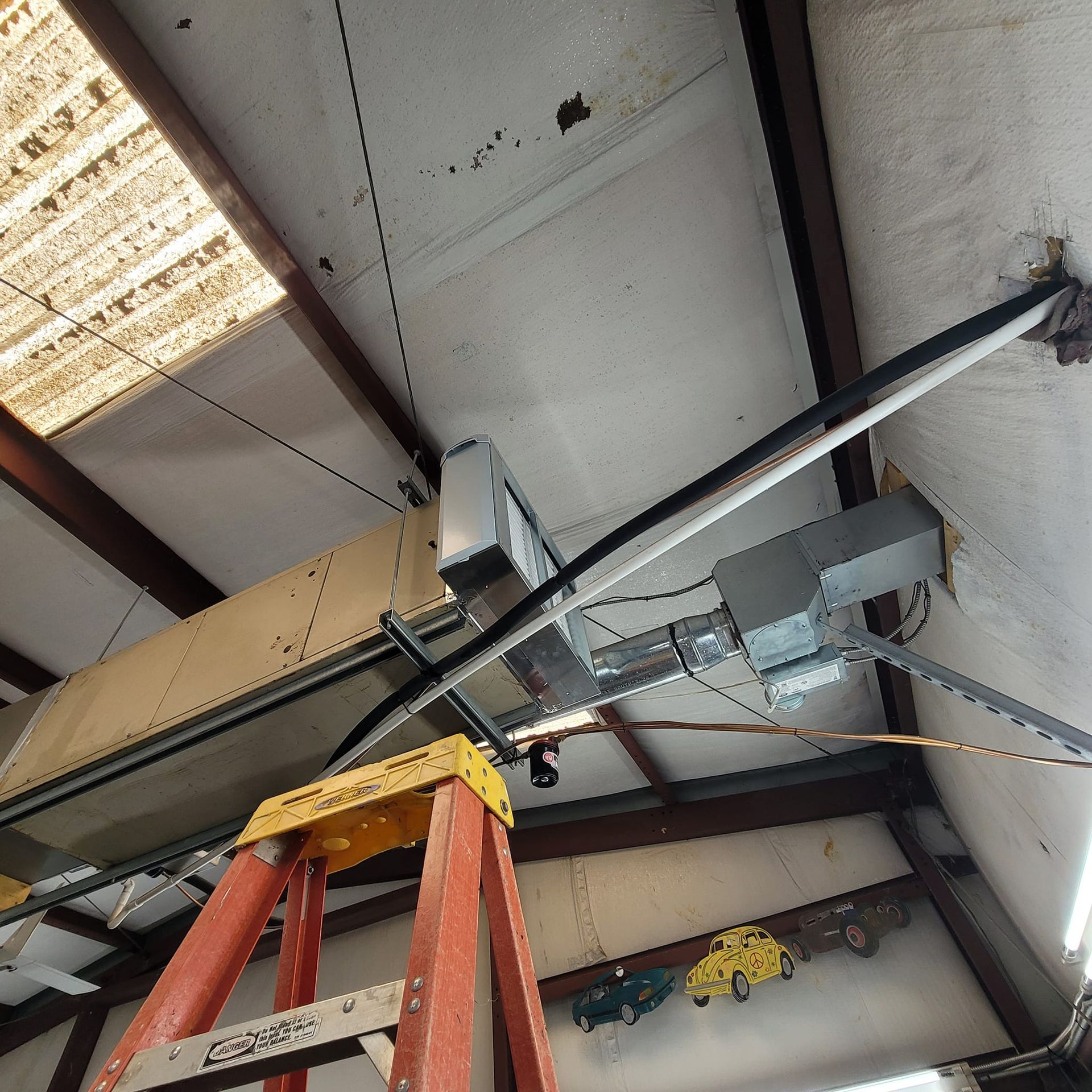 HVAC unit mounted to ceiling with ladder beneath; interior shot with car models on wall.