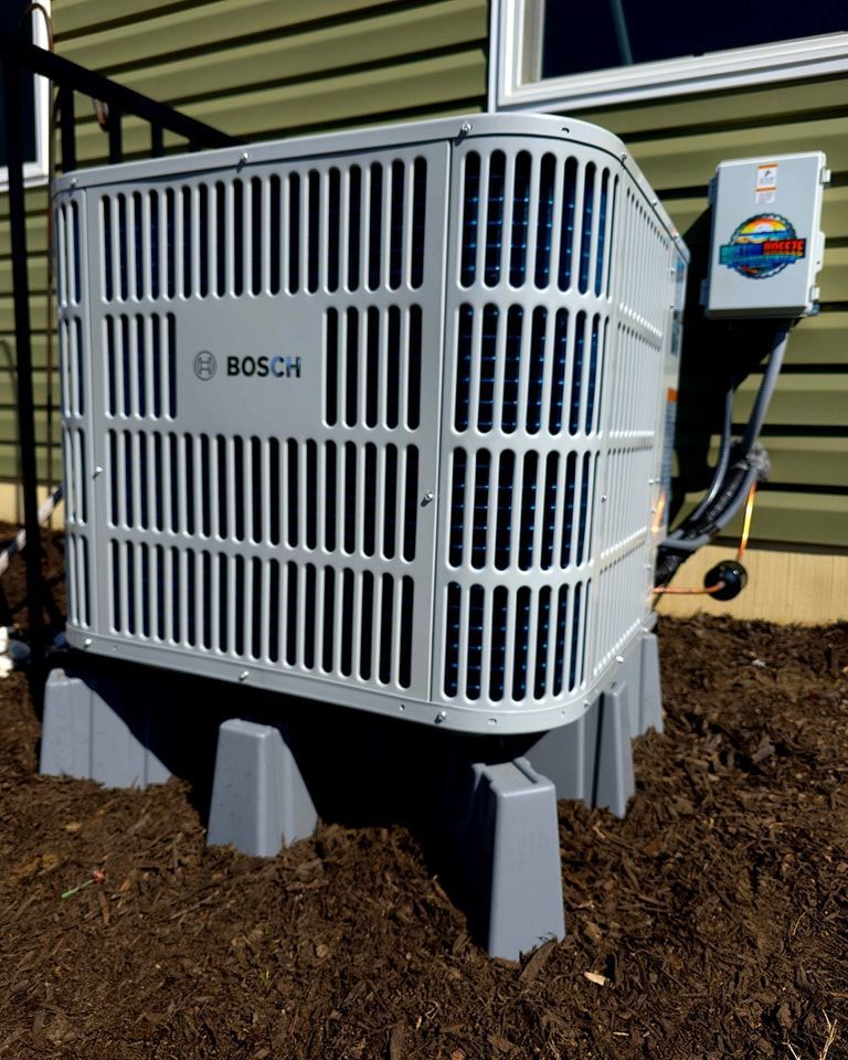 Bosch air conditioning unit on gray supports, next to a building with green siding and brown mulch.