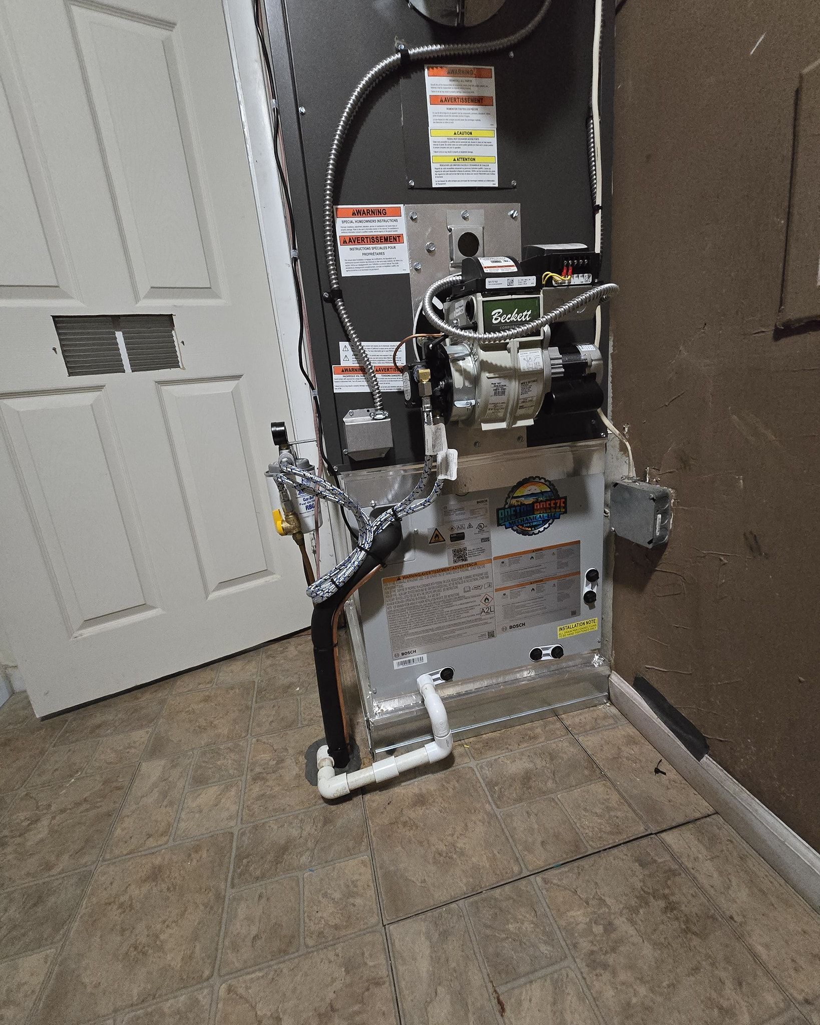 Furnace unit against a wall, next to a white door. Piping and wires are visible.